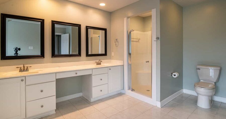 Spacious bathroom with a long white vanity, three black-framed mirrors, overhead recessed lighting, enclosed glass shower, tiled floor, and light gray walls