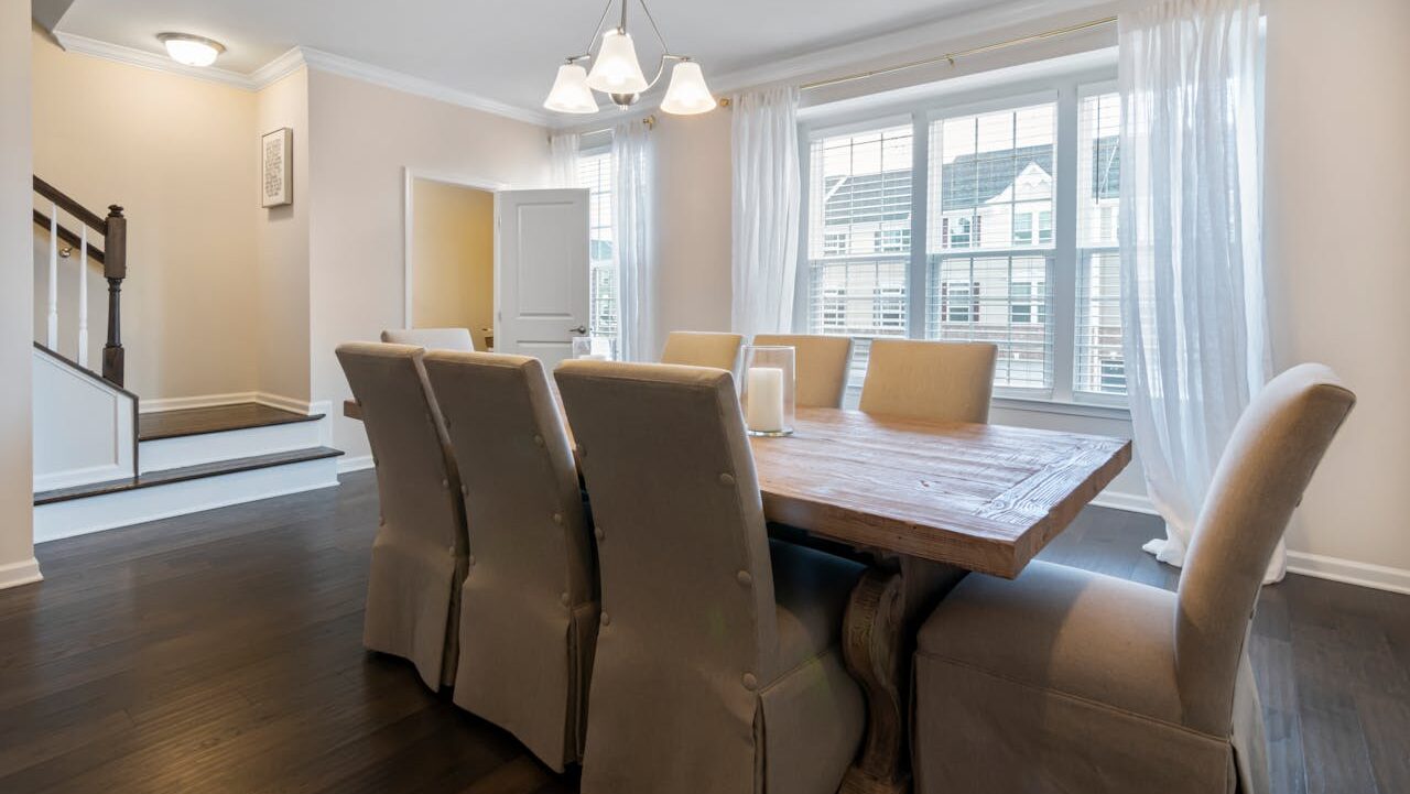 Dining room with a wooden table, beige chairs, and a simple light fixture, large windows with white curtains, staircase on the left, neutral-colored walls, a modern, tidy interior, a few candles on the table, cozy and clean atmosphere