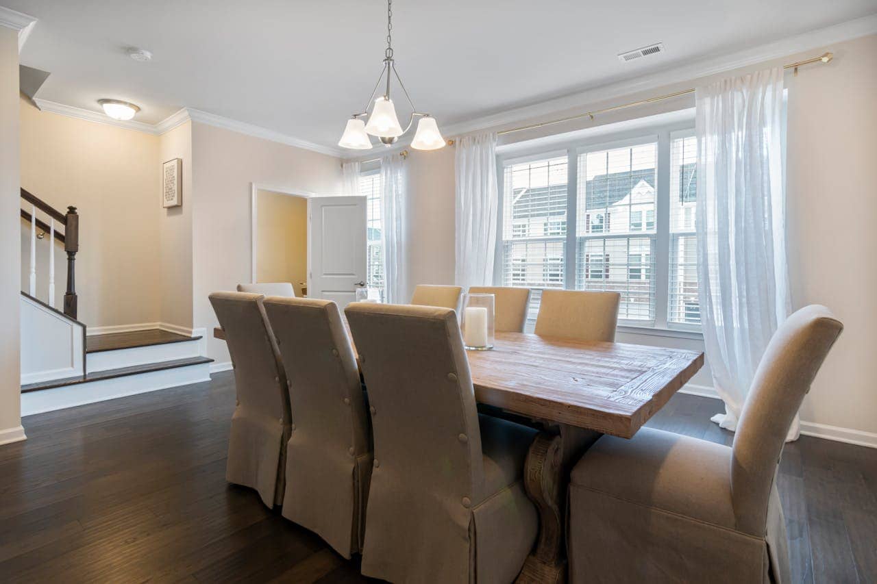 Dining room with a wooden table, beige chairs, and a simple light fixture, large windows with white curtains, staircase on the left, neutral-colored walls, a modern, tidy interior, a few candles on the table, cozy and clean atmosphere