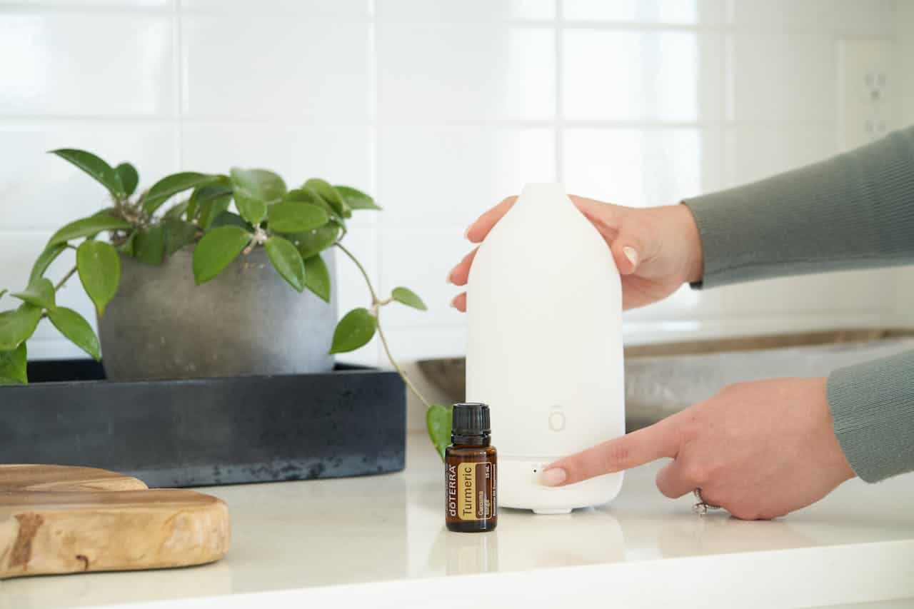 Person pressing a button on a white diffuser, plant in a gray pot on the counter, bottle of doTERRA turmeric essential oil next to the diffuser, clean modern kitchen setting