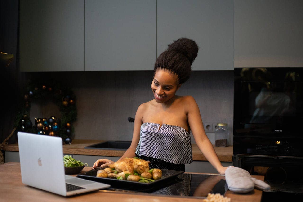 A woman in a kitchen smiling while looking at a tray of roasted chicken and vegetables, with a laptop placed in front of her on the counter, kitchen decor includes a Christmas wreath and an oven in the background