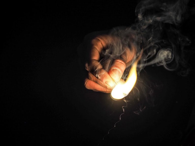 A hand holding a match, striking a flame, with smoke rising, dark background, close-up of fire ignition, lighting a match, creating a fire source, survival task, preparing for fire