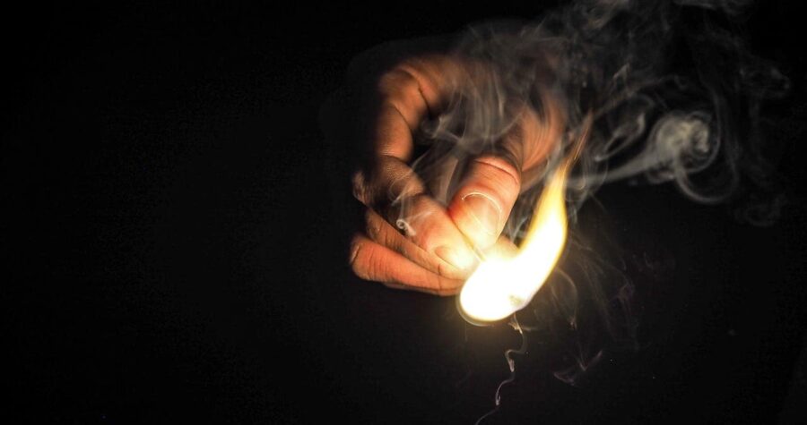 A hand holding a match, striking a flame, with smoke rising, dark background, close-up of fire ignition, lighting a match, creating a fire source, survival task, preparing for fire