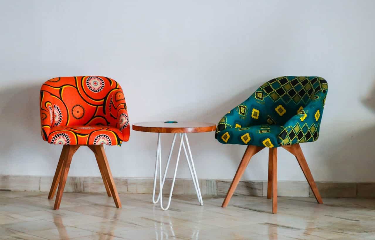 A pair of chairs with colorful, patterned fabric, one chair has an orange and yellow design, the other has a green and yellow pattern, a small round table sits between the chairs, with wooden legs and a white metal frame
