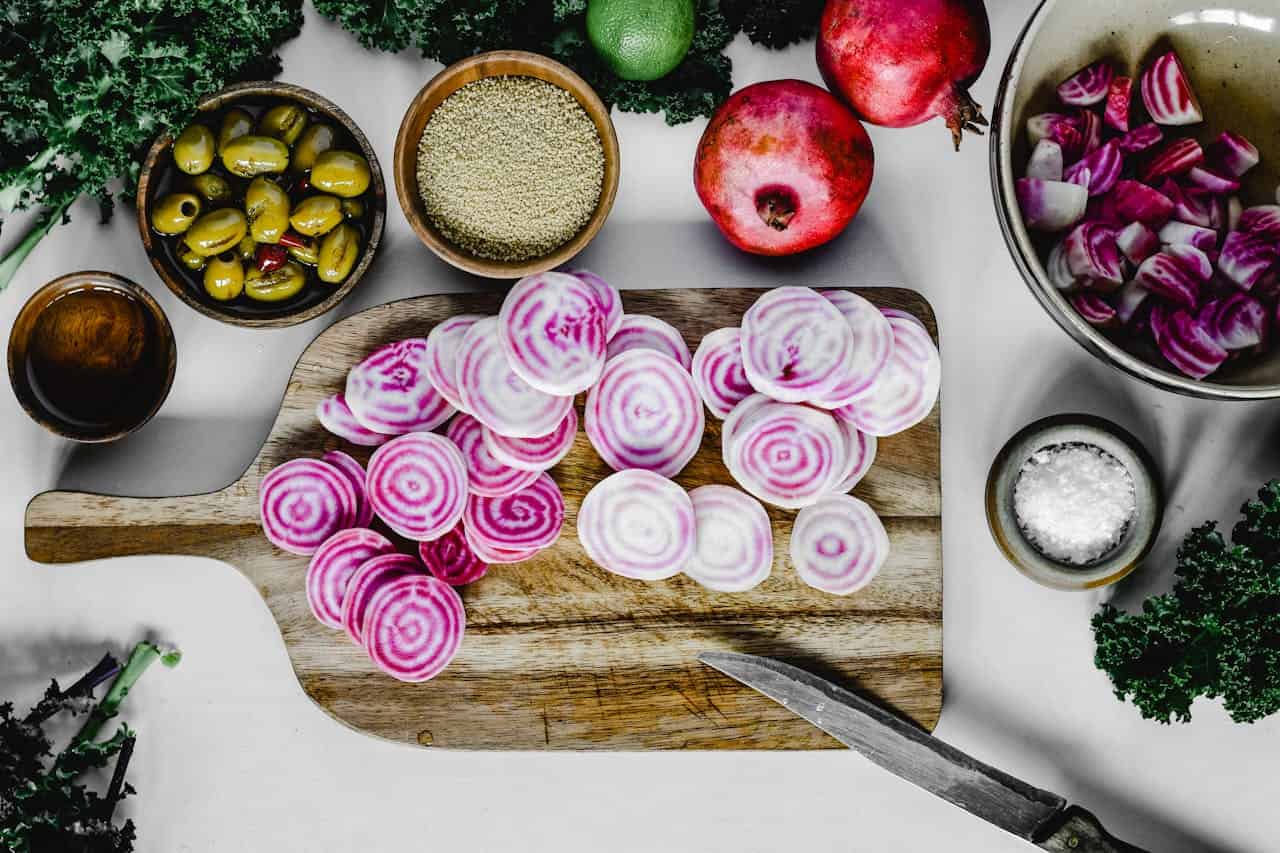 Sliced beetroot arranged on a wooden cutting board, bowls with green olives, quinoa, pomegranate, and chopped beetroot, salt in a small bowl