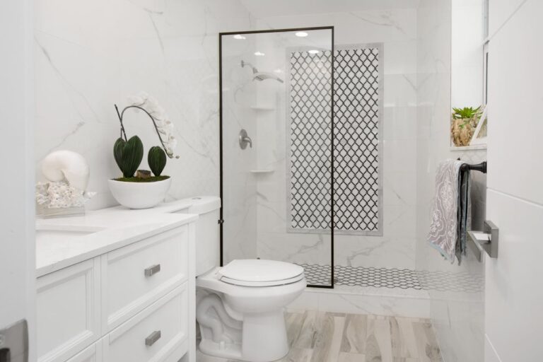 The image shows a modern, minimalist bathroom with a white and black theme, a clean design, a shower with a glass door and black accents, a toilet, a vanity with a countertop sink, a decorative plant and shells