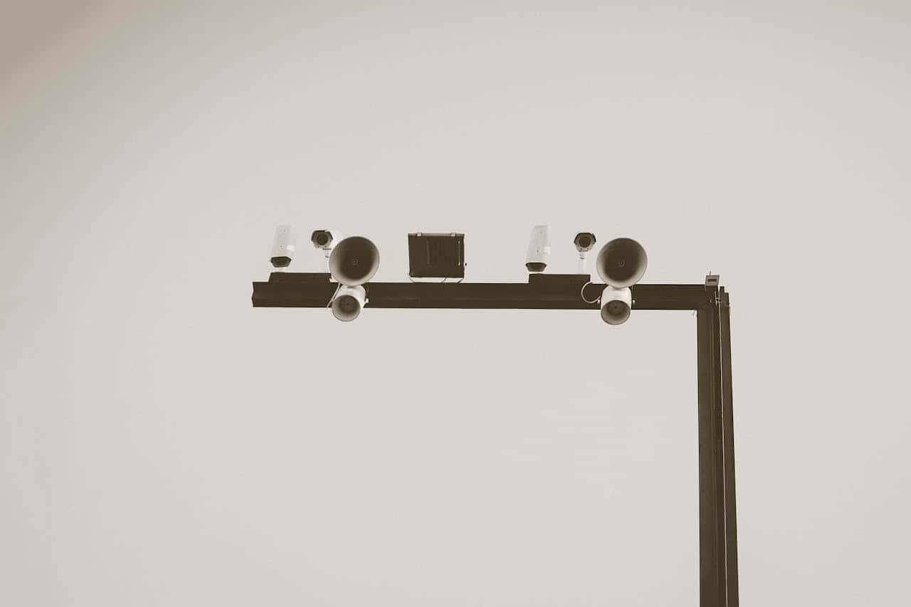 Surveillance camera system mounted on a metal frame, includes several cameras and speakers, monitoring or security setup, designed to track and broadcast, positioned overhead for wide coverage, black and white photo effect