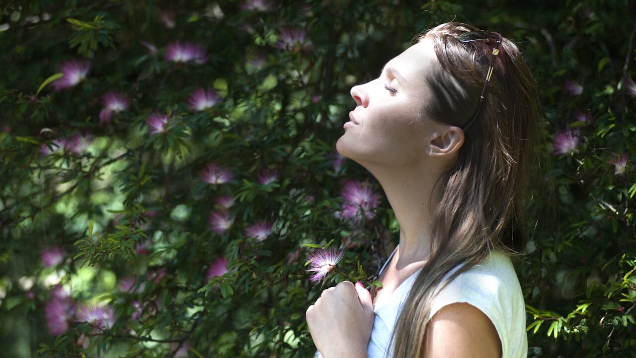 Woman enjoying nature, standing in a garden, looking up, eyes closed, taking a deep breath, peaceful expression, surrounded by green plants, pink flowers in the background, relaxing moment