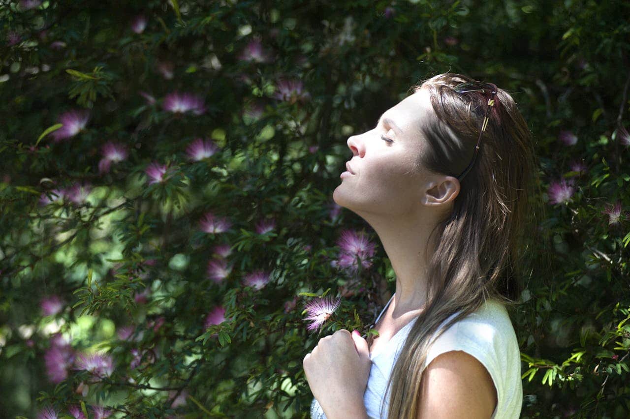 Woman enjoying nature, standing in a garden, looking up, eyes closed, taking a deep breath, peaceful expression, surrounded by green plants, pink flowers in the background, relaxing moment