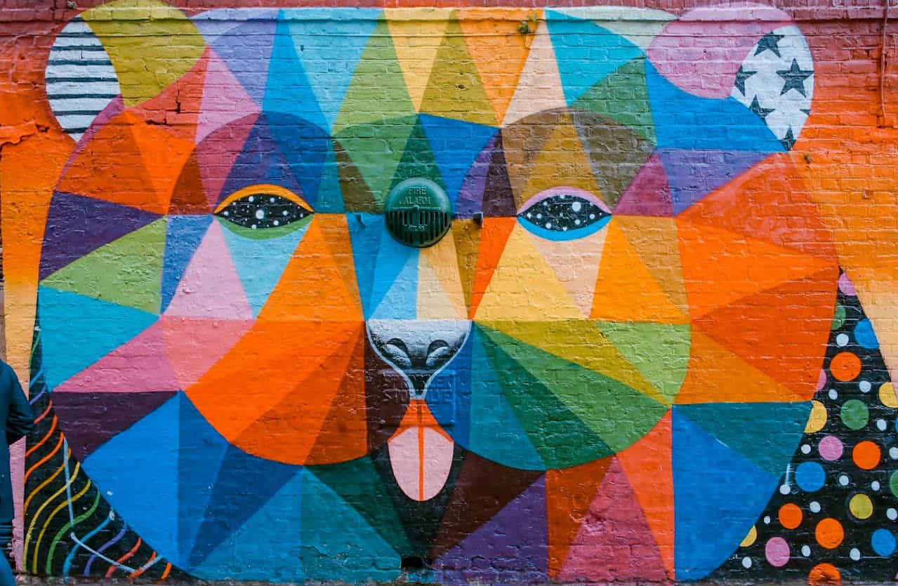 A vibrant, colorful mural of a bear’s face made of geometric shapes, featuring bold oranges, blues, greens, and purples, with stars and dots in the eyes and surrounding patterns