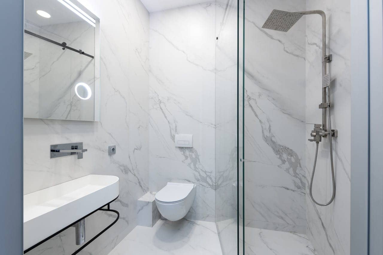 Modern bathroom with sleek white marble walls, a minimalist wall-mounted toilet, a shower with glass enclosure and overhead showerhead, a contemporary sink with a mirror featuring integrated lighting, simple and elegant design emphasizing clean lines and space efficiency