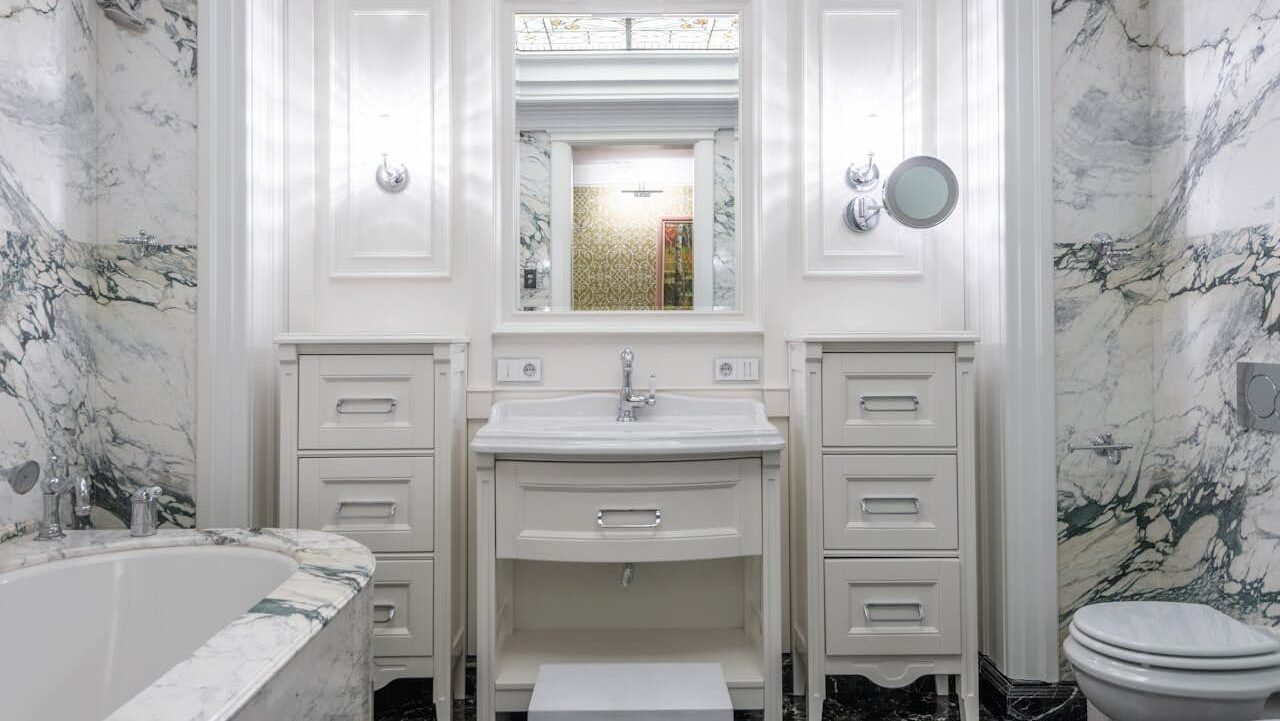 Elegant white marble bathroom with vanity sink, cabinet drawers, bathtub, toilet, mirror, and classical architectural details