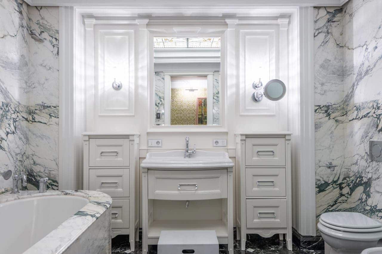 Elegant white marble bathroom with vanity sink, cabinet drawers, bathtub, toilet, mirror, and classical architectural details