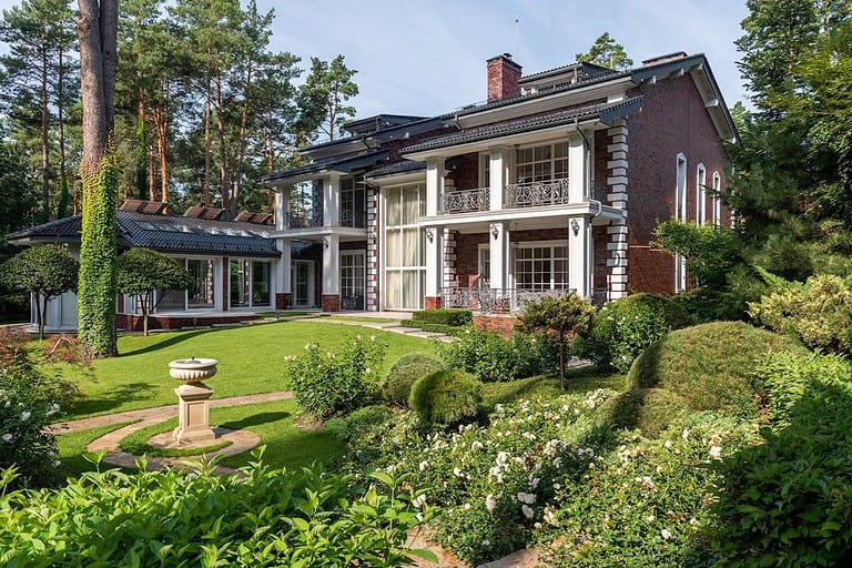 Elegant two-story brick mansion with white columns, manicured lawn, lush garden, wrought iron balconies, and surrounded by tall pine trees