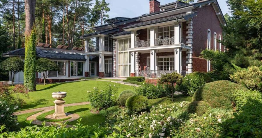 Elegant two-story brick mansion with white columns, manicured lawn, lush garden, wrought iron balconies, and surrounded by tall pine trees