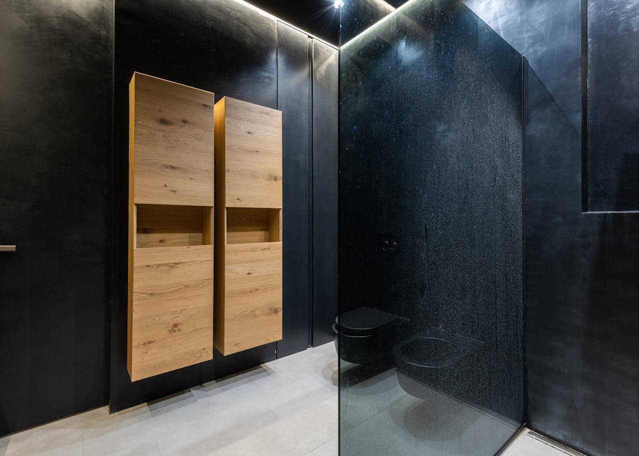 Modern bathroom design with dark black walls, wooden storage units with open shelves, sleek black toilet and shower area, minimalistic decor, shower glass with water droplets, clean and contemporary aesthetic, well-lit with subtle overhead lighting