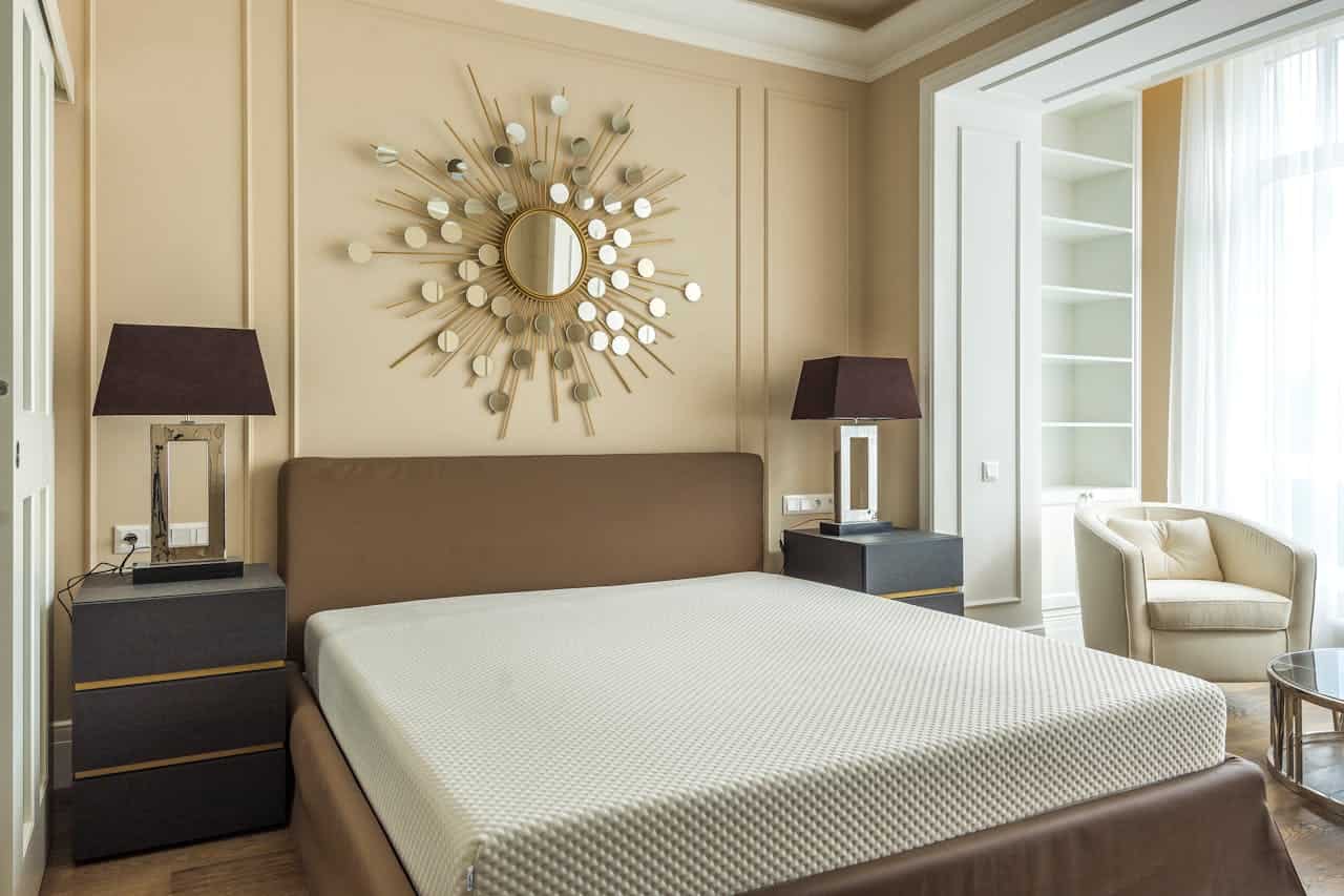 Modern bedroom with a brown bed, abstract sunburst wall mirror, two lamps, and a cozy chair by the window