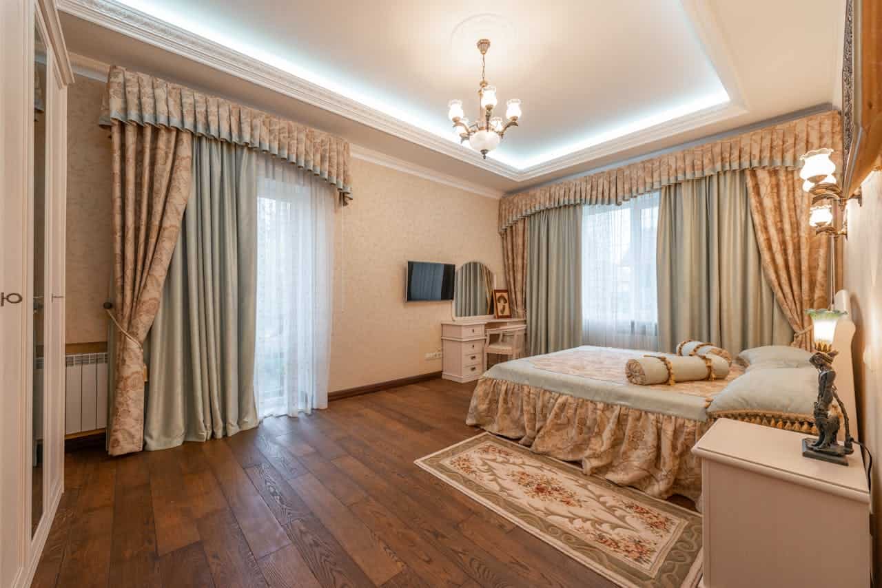 A well-lit bedroom with elegant decor, light blue and beige curtains framing a window, a plush bed with matching pillows and a decorative bed skirt, a small side table with a lamp and a statue, a TV mounted on the wall, wooden flooring and soft lighting creating a cozy, comfortable atmosphere