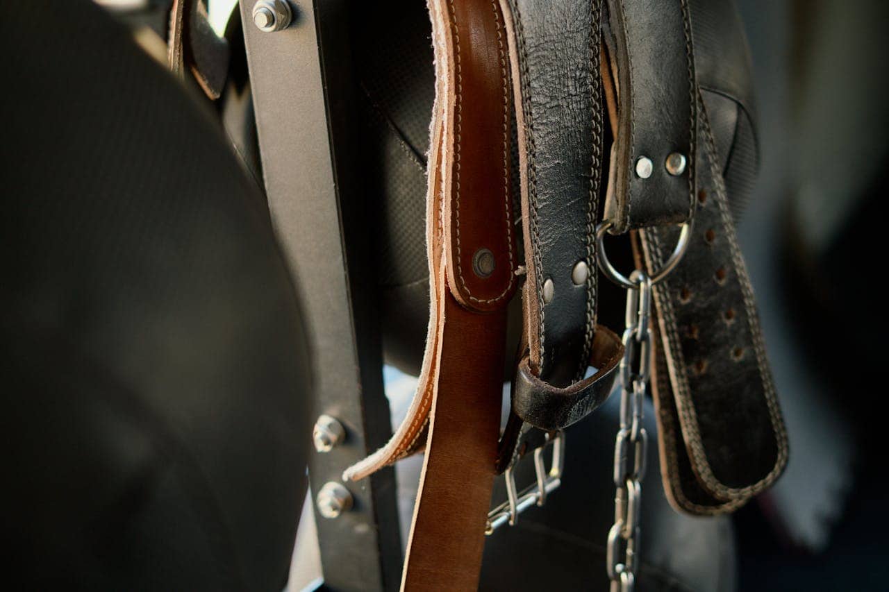 Leather belts hanging on a rack, with one belt held by a metal chain, close-up view of textured leather, metal studs and holes visible on the belts, dark background with a focus on the belts, neat organization of accessories