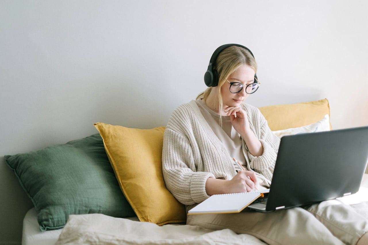 A person sitting on a bed, wearing headphones, writing in a notebook, working on a laptop, wearing a beige cardigan, with yellow and green pillows around them, calm and focused environment, relaxed atmosphere for studying or working at home, indoor setting with natural light