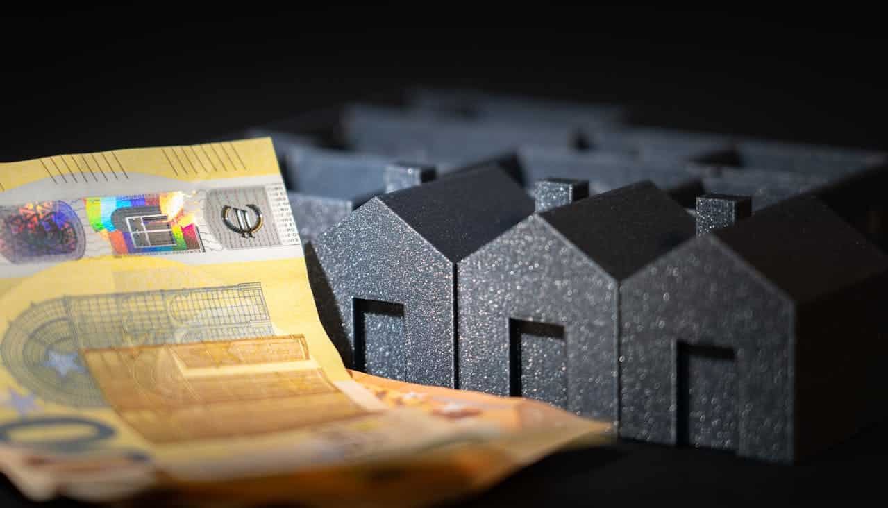 A close-up of a banknote and a model of small, black, glittery houses in the background, symbolizing real estate investment and market value