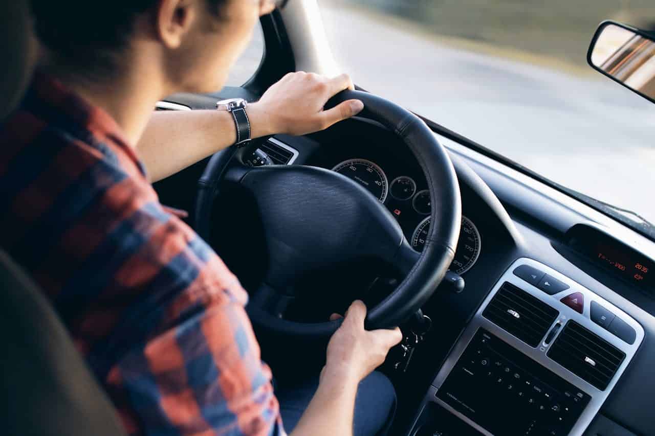 A person driving a car, holding the steering wheel with both hands, focused on the road ahead, interior view of the car showing the dashboard and controls