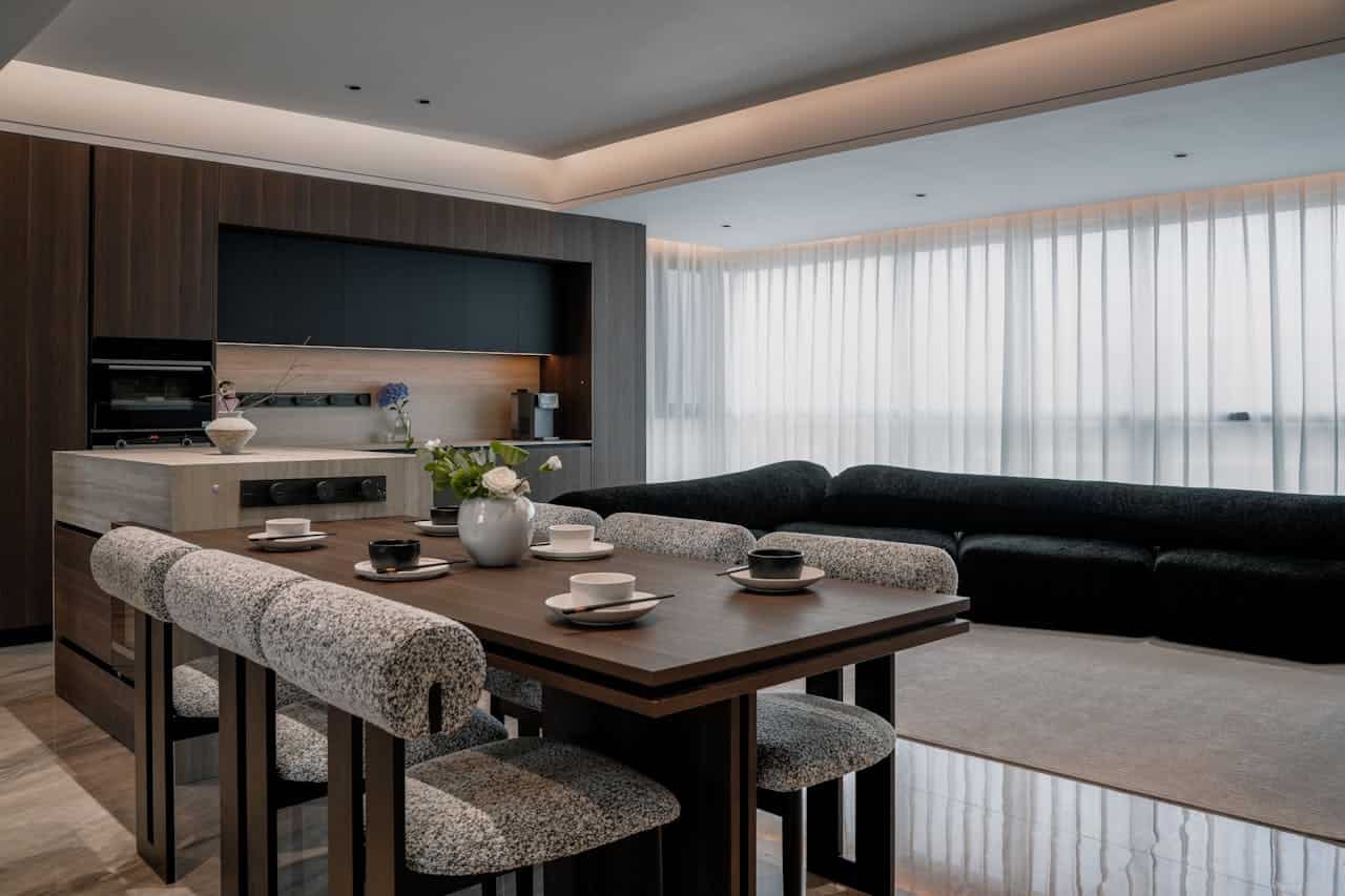 Modern kitchen with sleek dark wood cabinetry, large island with stove, minimalistic white vase with flowers, comfortable dining area with two-tone upholstered chairs