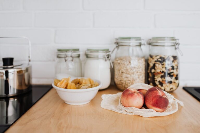 Wooden kitchen counter with glass storage jars, a bowl of chips, peaches on mesh cloth, and kettle against white wall