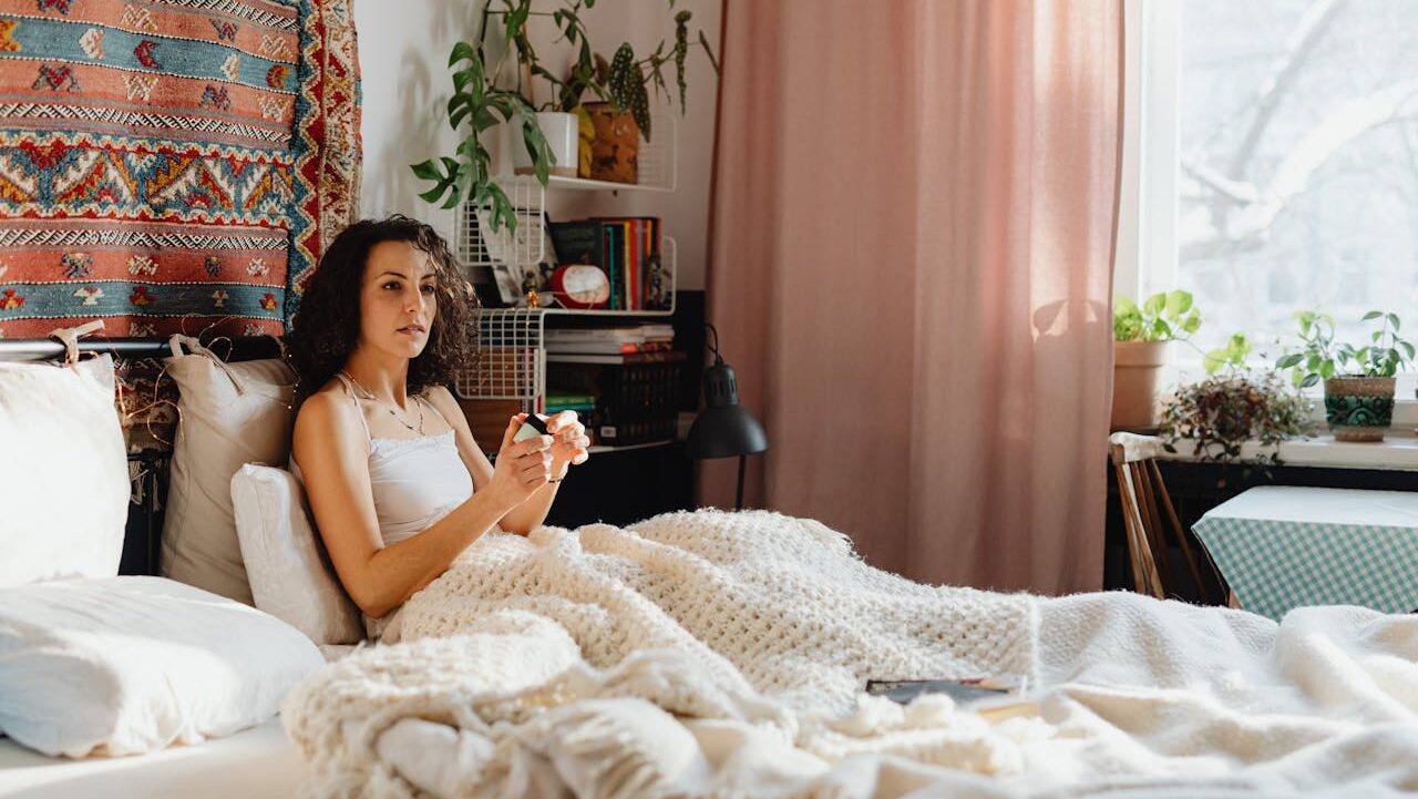 A person sitting on a bed wrapped in a blanket, holding an object in their hand, relaxed expression, surrounded by pillows, natural light from a window, plants in the background, cozy and calm room with soft curtains, casual and comfortable setting