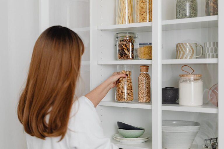 A woman placing a jar filled with seeds on a white pantry shelf, other jars containing dried mushrooms, pasta, and other items, a set of ceramic bowls and cups on the lower shelf, a clean, organized pantry