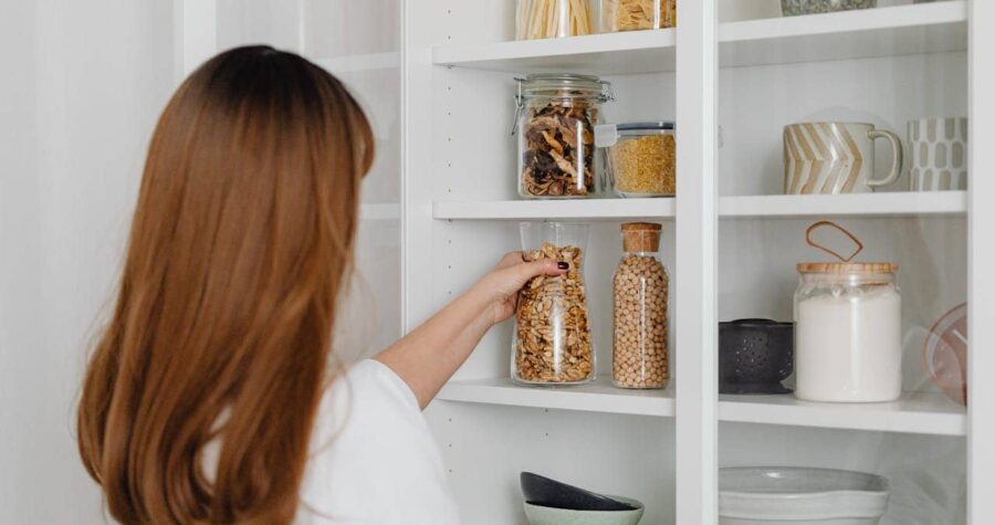 A woman placing a jar filled with seeds on a white pantry shelf, other jars containing dried mushrooms, pasta, and other items, a set of ceramic bowls and cups on the lower shelf, a clean, organized pantry