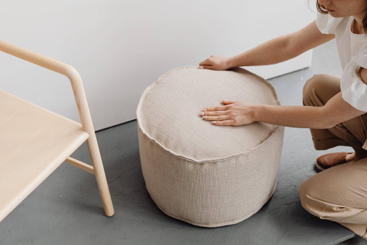 A person adjusting a beige fabric pouf, a wooden chair is beside it, light-colored floor, modern, minimalistic interior, focus on the hand pressing the top of the pouf, soft lighting, casual home setting