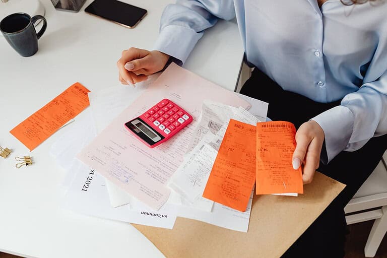 A person organizing receipts, using a pink calculator, sitting at a desk with papers and receipts, holding orange receipts in one hand, surrounded by various documents, coffee cup and phone visible in the background, paper clips nearby, indicating work or personal accounting tasks