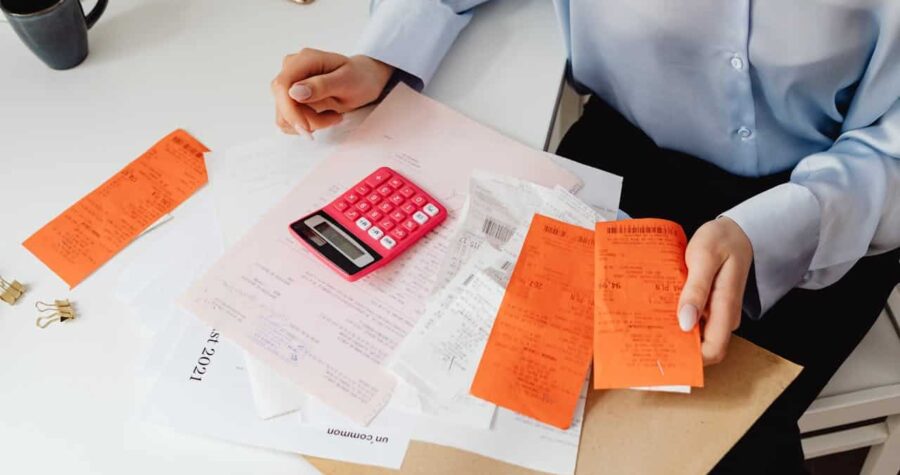 A person organizing receipts, using a pink calculator, sitting at a desk with papers and receipts, holding orange receipts in one hand, surrounded by various documents, coffee cup and phone visible in the background, paper clips nearby, indicating work or personal accounting tasks