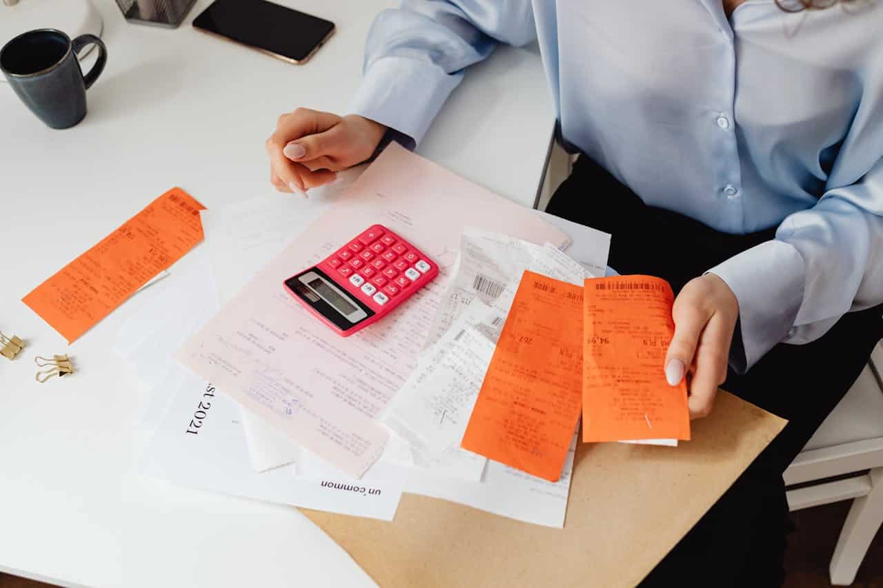 A person organizing receipts, using a pink calculator, sitting at a desk with papers and receipts, holding orange receipts in one hand, surrounded by various documents, coffee cup and phone visible in the background, paper clips nearby, indicating work or personal accounting tasks