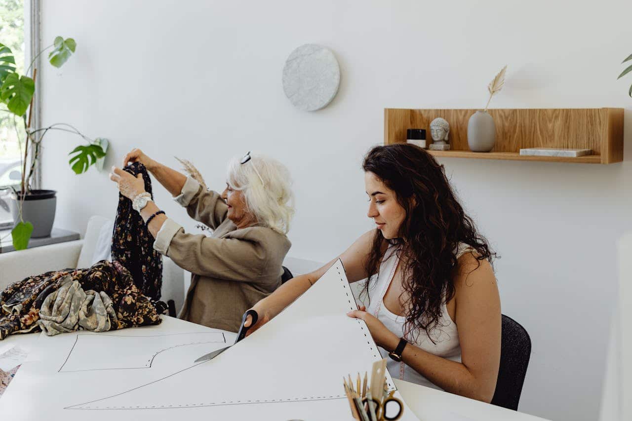 A woman cutting fabric with scissors, working on a pattern, while another person folds clothing, in a bright, modern workspace, focused on design or sewing,
