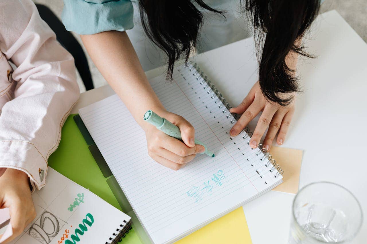 A person writing with a green marker on a notebook, hand resting on a spiral-bound notebook with lined pages, colorful papers and a glass of water on the table