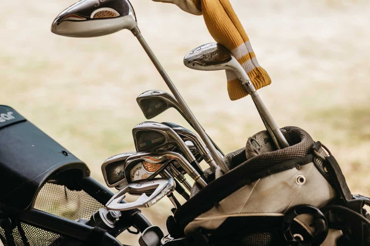 Golf clubs in a bag, driver and iron clubs, golf bag with shoulder strap, golf gloves hanging, outdoors, preparing for a game, set of golf equipment