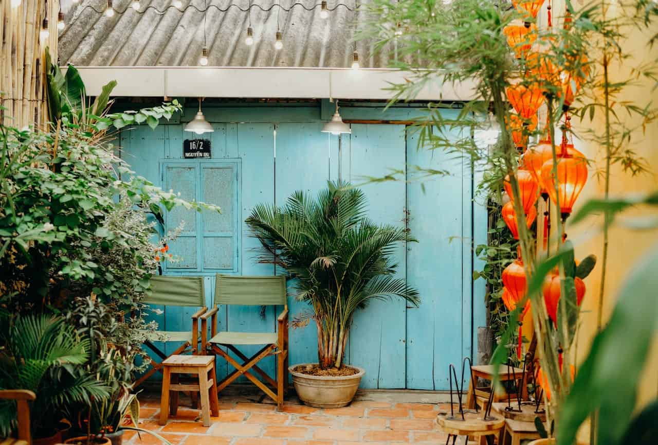 A cozy outdoor seating area with two green chairs and a small wooden stool, a blue wooden door with a sign, potted plants surrounding the space, colorful orange lanterns hanging on the right