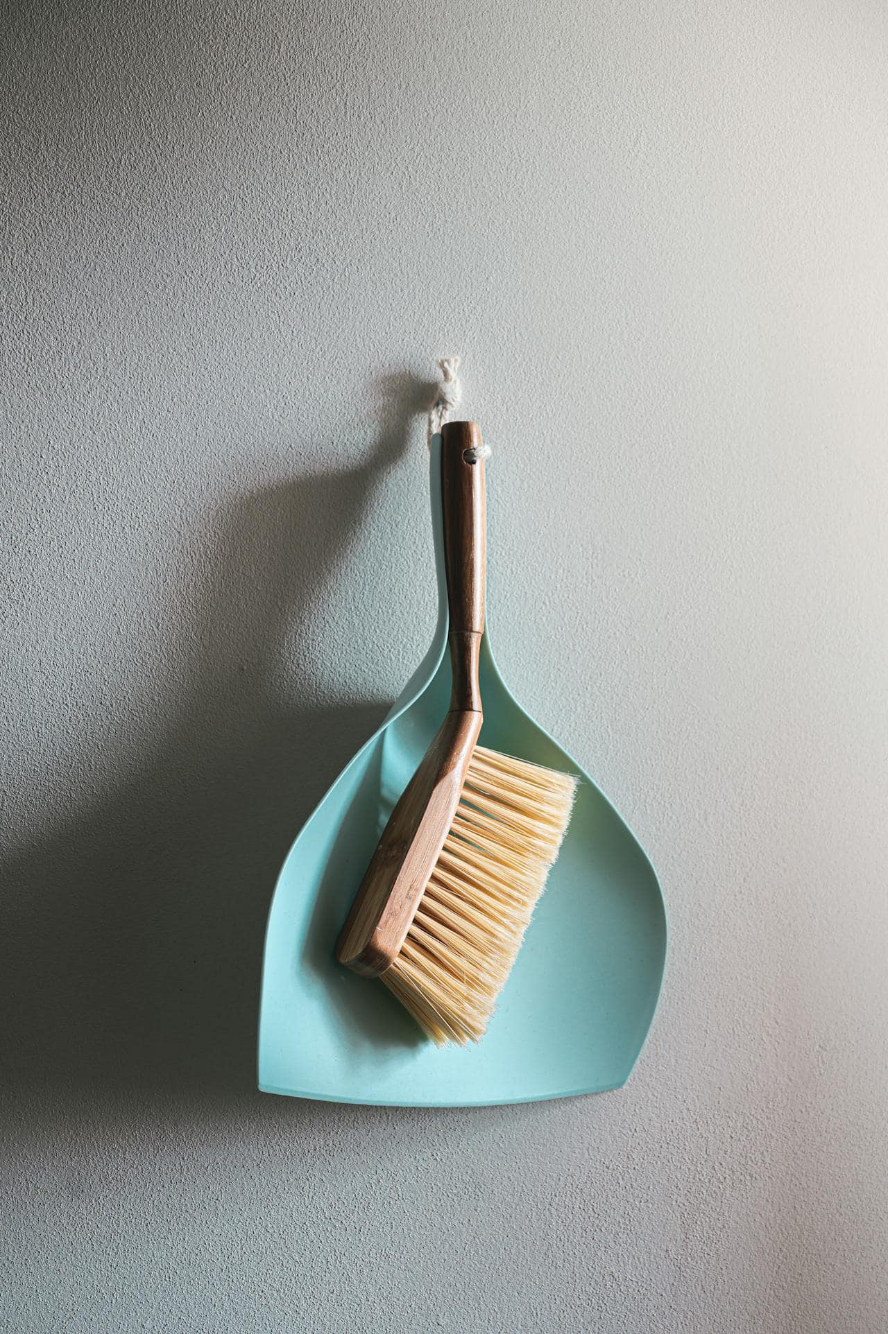 A light blue dustpan and a wooden-handled brush with beige bristles, hanging on a textured light gray wall