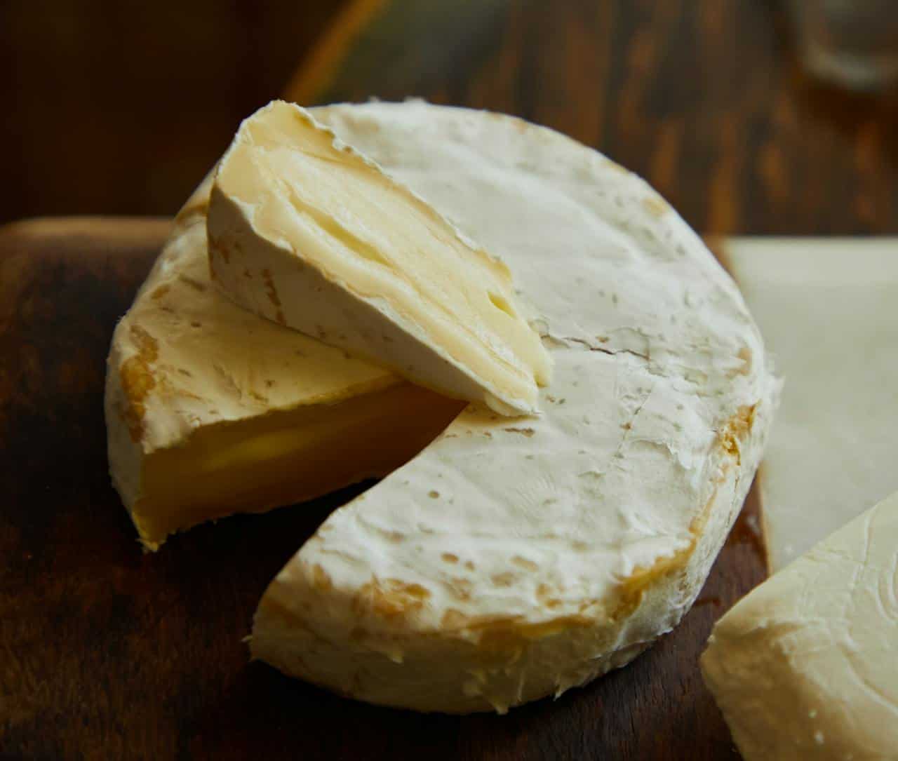 A wheel of soft cheese with a creamy interior is shown, a wedge has been cut and is partially removed, the cheese is wrapped in a white rind, placed on a wooden surface, showcasing its rich texture and smooth appearance
