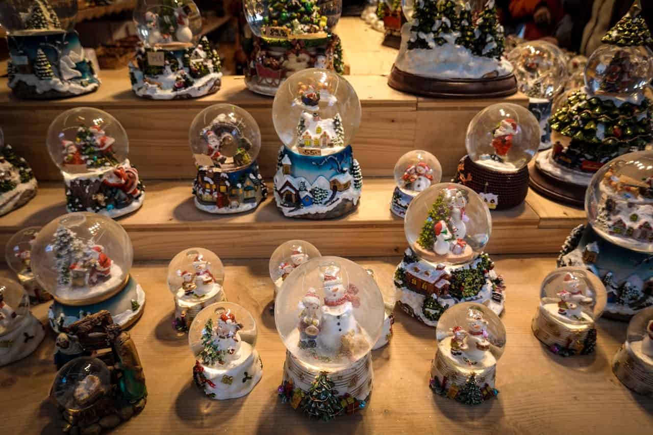 The image depicts multiple snow globes, each containing miniature Christmas-themed scenes like snowmen, Santa Claus, and festive houses, arranged in rows on a wooden surface