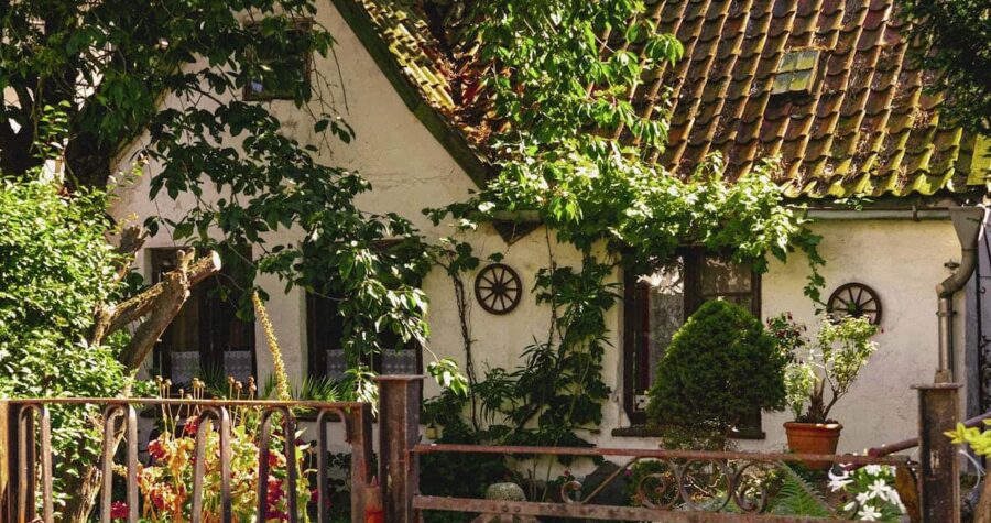 Charming white cottage with a rustic roof, surrounded by greenery and plants, decorative windows with round frames, a wrought iron gate in front, garden flowers, potted plants, peaceful and cozy atmosphere