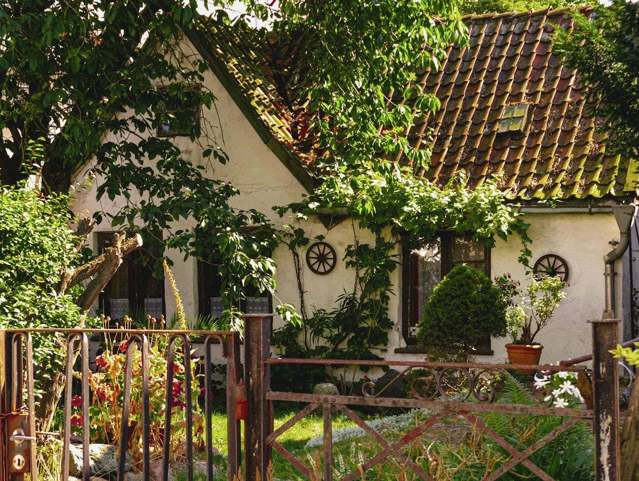 Charming white cottage with a rustic roof, surrounded by greenery and plants, decorative windows with round frames, a wrought iron gate in front, garden flowers, potted plants, peaceful and cozy atmosphere