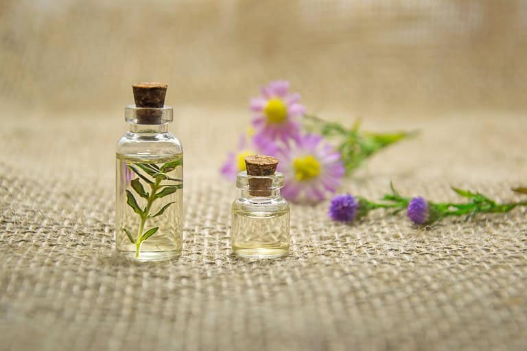 Two glass bottles with cork stoppers, containing essential oil and a small plant inside, placed on a burlap surface, with purple and yellow flowers in the background, essential oils