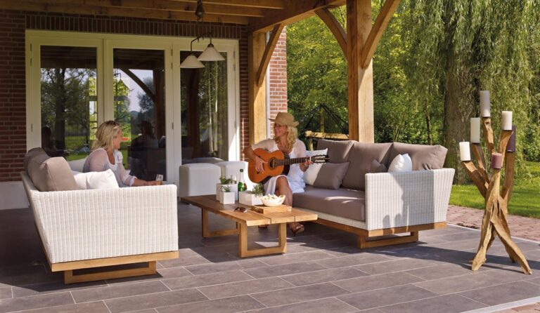 Two women relaxing on a patio, one sitting and enjoying a drink, the other playing guitar, a wooden table with drinks and snacks between them, comfortable wicker sofas, soft cushions