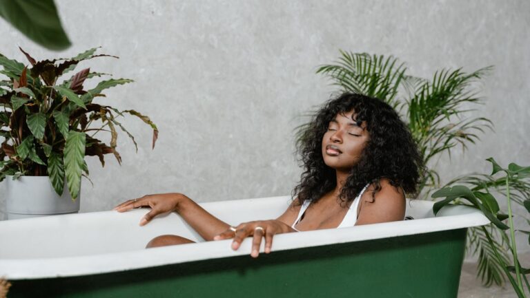 A woman with curly hair relaxes in a green bathtub, surrounded by plants, her eyes are closed in a peaceful expression, she is wearing a white top, and the bathroom has a neutral gray wall background with a clean, minimalist aesthetic