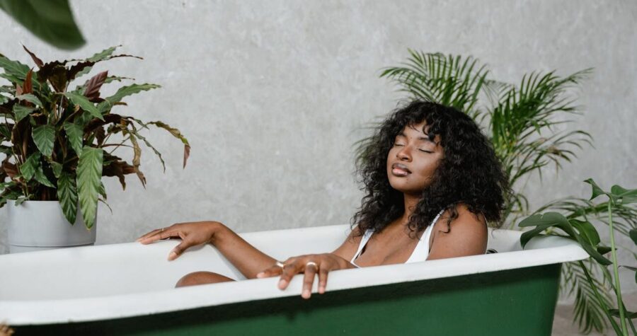 A woman with curly hair relaxes in a green bathtub, surrounded by plants, her eyes are closed in a peaceful expression, she is wearing a white top, and the bathroom has a neutral gray wall background with a clean, minimalist aesthetic