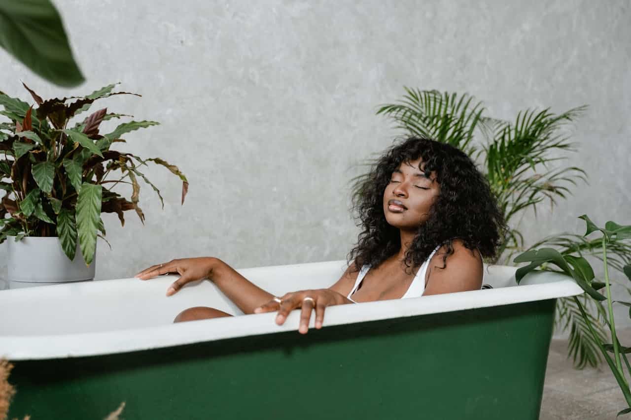 A woman with curly hair relaxes in a green bathtub, surrounded by plants, her eyes are closed in a peaceful expression, she is wearing a white top, and the bathroom has a neutral gray wall background with a clean, minimalist aesthetic