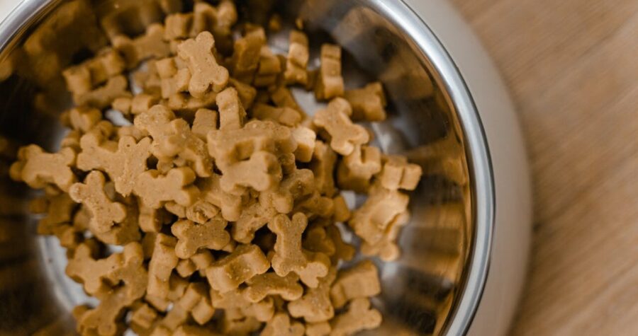 Metal bowl filled with small, bone-shaped dog treats, placed on a wooden surface, treats are light brown and uniform in size, likely prepared for feeding a pet
